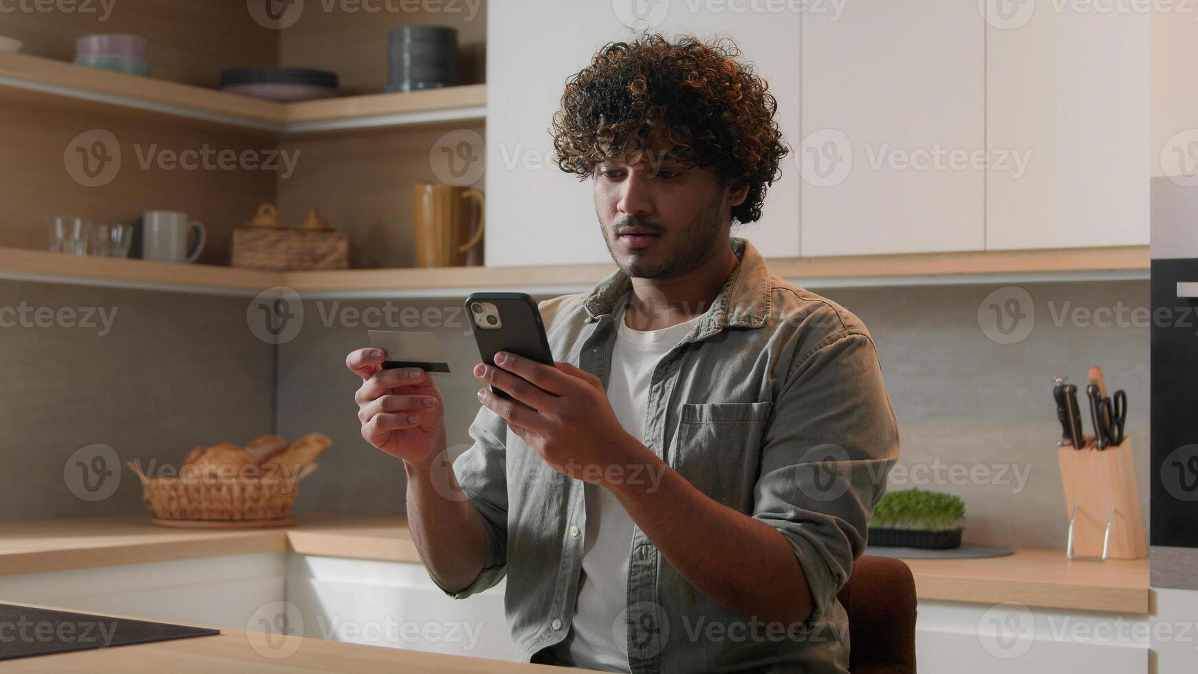 Indian male customer man Arabian guy homeowner sitting at kitchen order food products with smart phone app using bank credit card making purchases at web grocery store delivery service shopping online photo