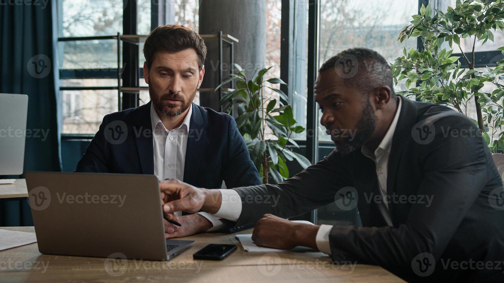 Two diverse multiracial businessmen discuss online data project with laptop in office writing notes documents. Caucasian businessman and African American man colleagues men company working together photo