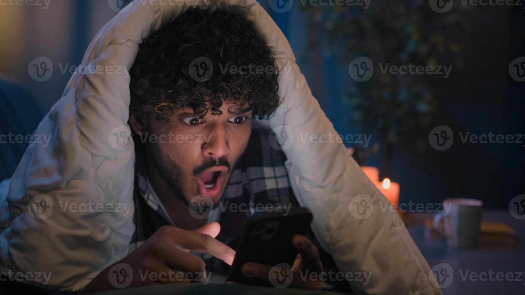 Shocked guy Indian man Arabian male shock wonder reading news in phone social media amazed with smartphone message at night evening home on couch under blanket cover with duvet surprised gadget addict photo