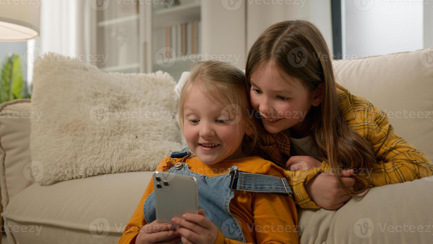 Two funny happy caucasian kids girls sisters friends children having fun playing on phone in living room couch play mobile online game together using smartphone browsing social network in Internet photo