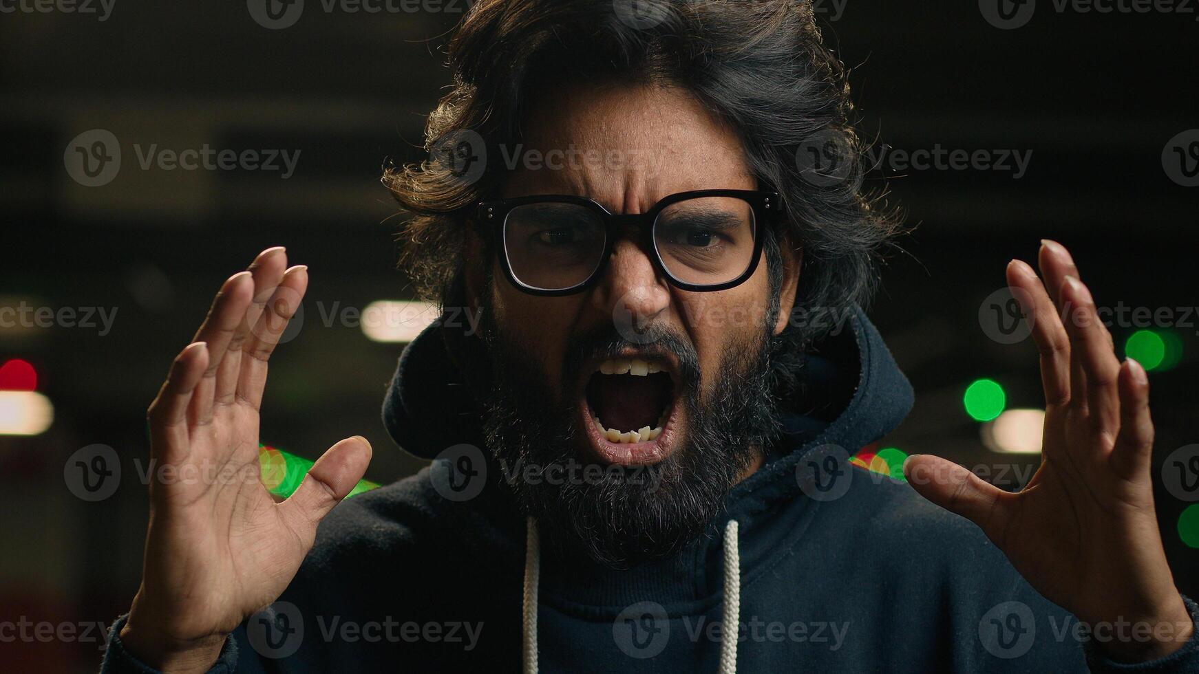 Close up angry mad aggressive Arabian bearded man in glasses scream at dark parking portrait furious loud Indian male guy screaming shouting yelling at underground irritated shout stress anger rage photo