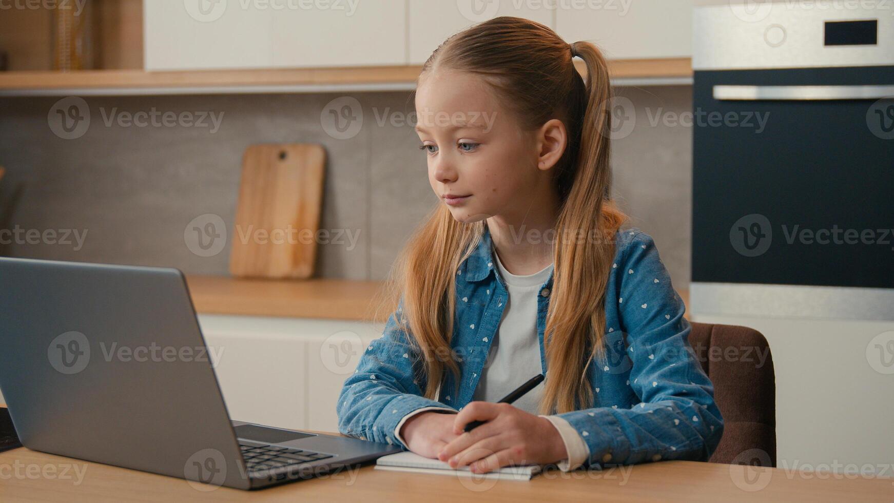 Caucasian kid girl schoolgirl listen online lesson school class distant teacher writing at home. Child pupil studying remote education with laptop call e-learning homework write task at kitchen photo