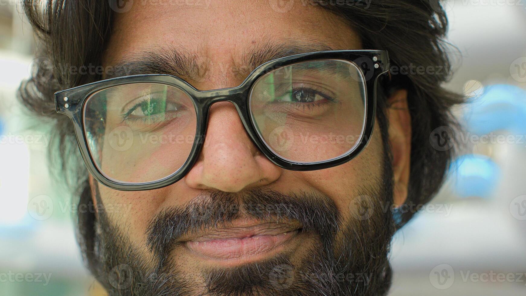 Close up positive male face in eyeglasses. Portrait Indian bearded man in glasses smiling looking at camera. Headshot entrepreneur guy businessman executive indoor with lights reflection in spectacles photo