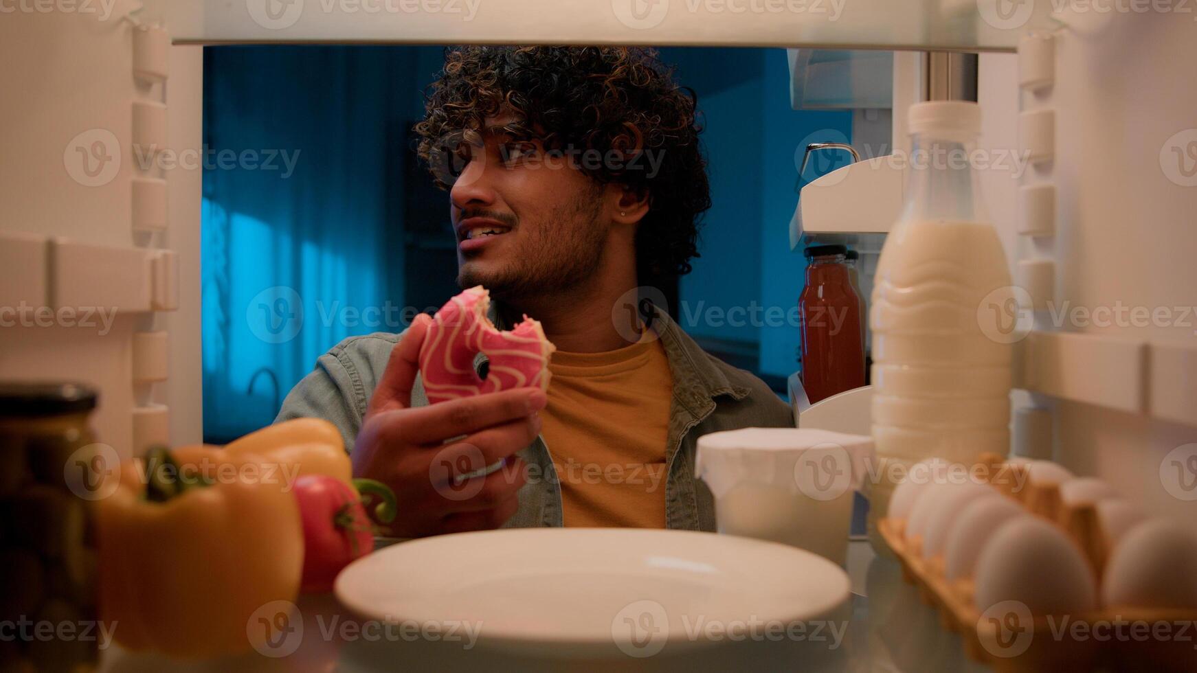 Point of view POV from inside refrigerator hungry Indian man Arabian guy male at night kitchen open fridge want eat take bitten sweet donut smiling condemn bakery unhealthy food nutrition overeating photo