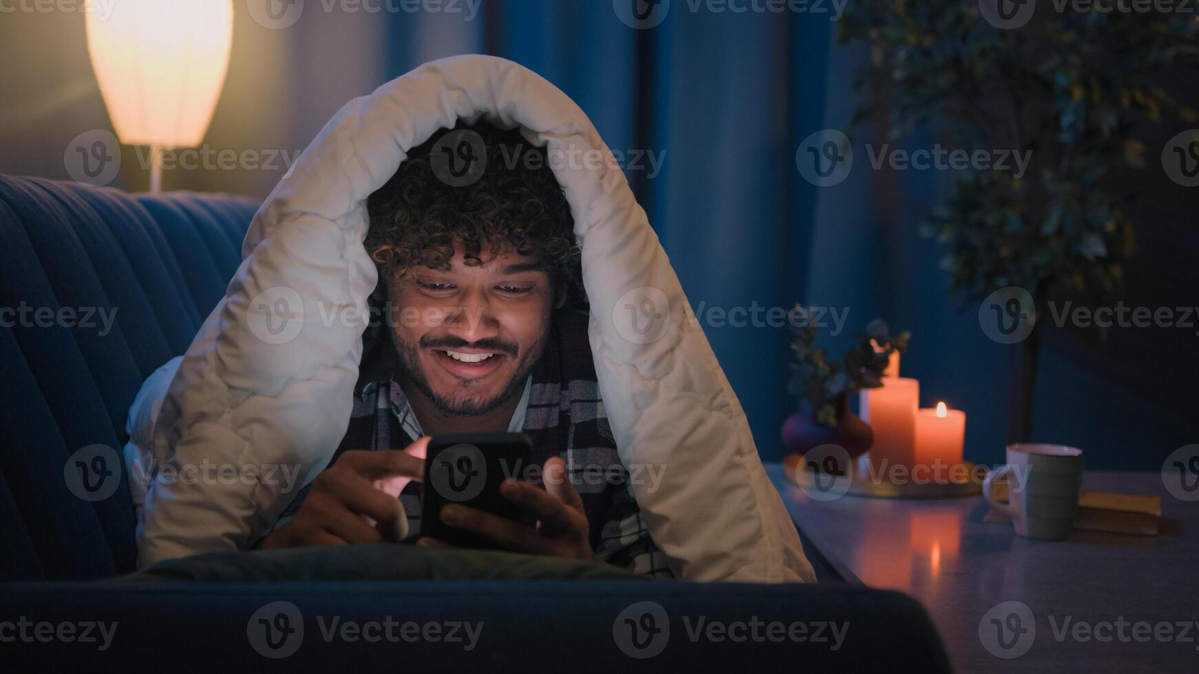 Smiling guy Indian man Arabian male at night evening home on couch under blanket cover with duvet looking mobile phone wow surprised amazed reading news gadget addict smartphone social media scrolling photo