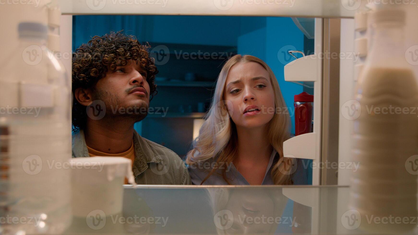Point of view POV inside refrigerator Caucasian girl woman girlfriend wife and Arabian man Indian guy boyfriend husband multiracial couple at night evening open empty fridge at kitchen unhappy sad photo