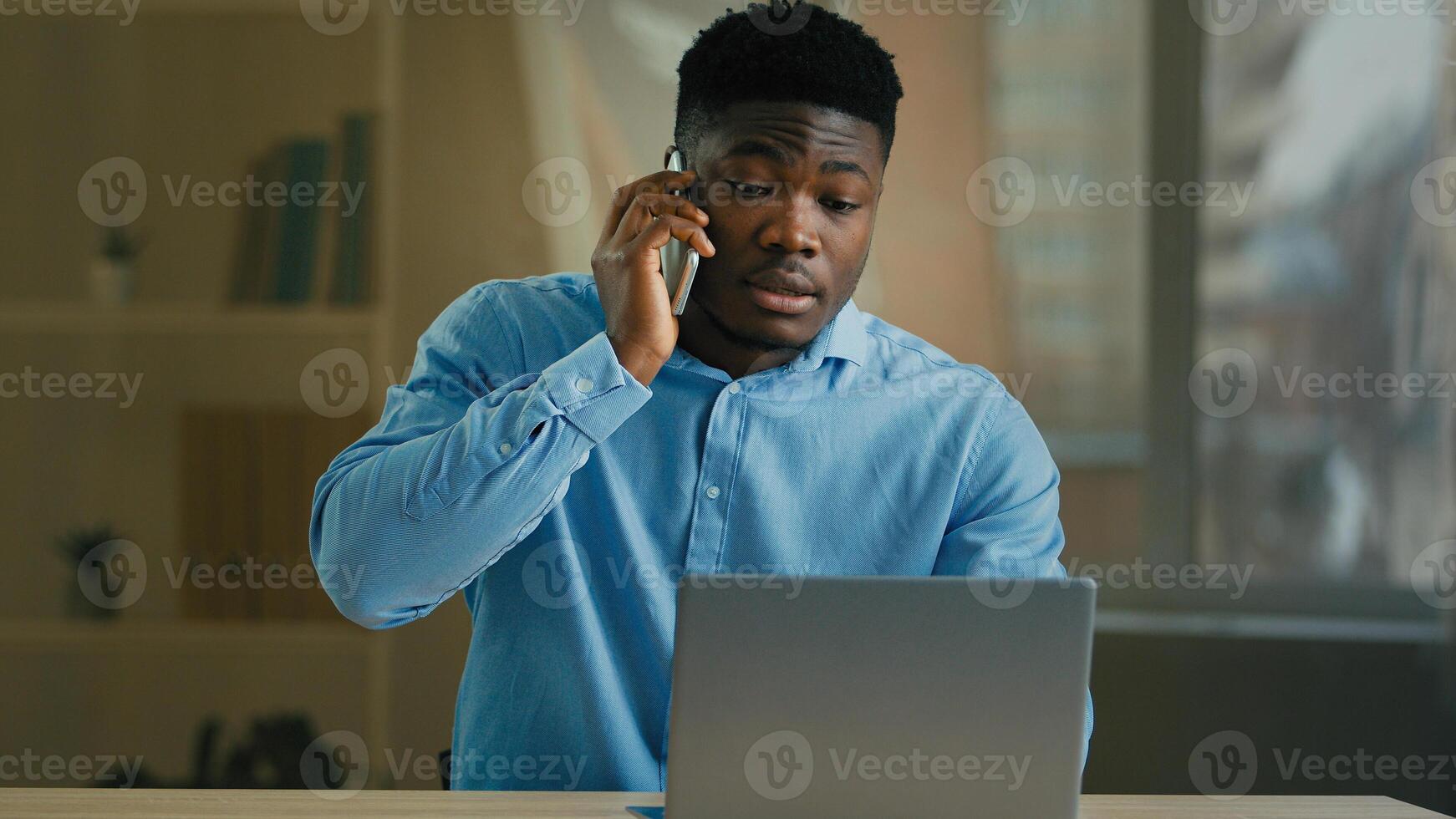 African male businessman ethnic 30s man office worker work distant from home talk business conversation by mobile phone wireless call discussing project look at laptop screen use cellphone smartphone photo