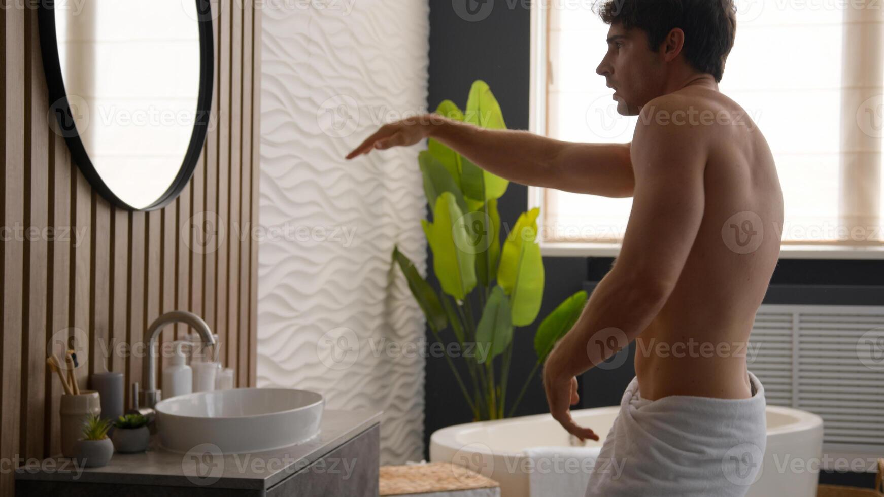 Funny Caucasian naked happy joyful male singing song joy man in bathroom with bath towel on hips dancing enjoying morning hygiene routine near mirror having fun dance listen music spinning around sing photo