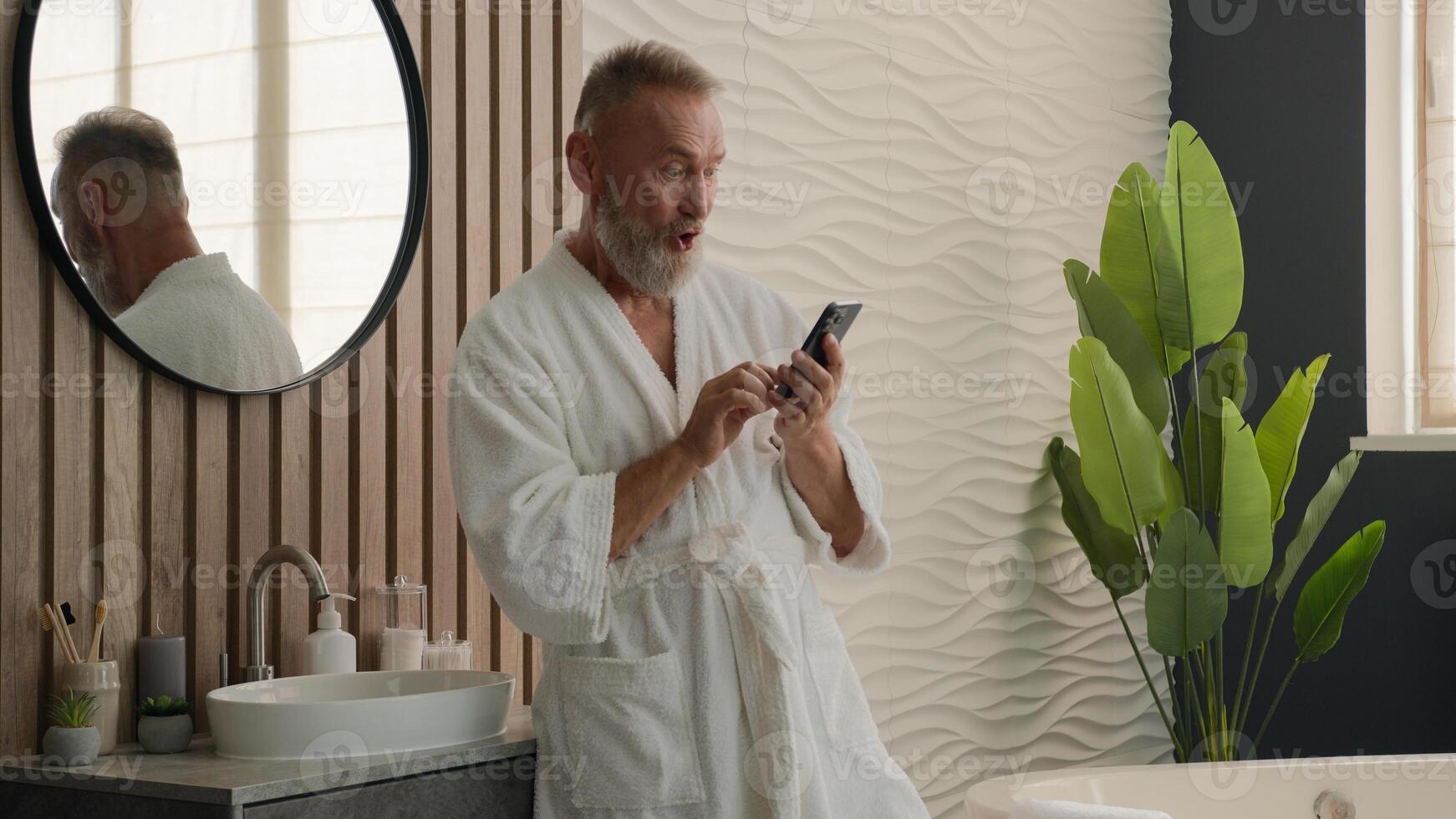 Old Caucasian man senior mature retired male in bathrobe in bath bathroom at morning looking mobile phone reading shocked good news amazed wonder wow reaction surprise smartphone sms message smiling photo