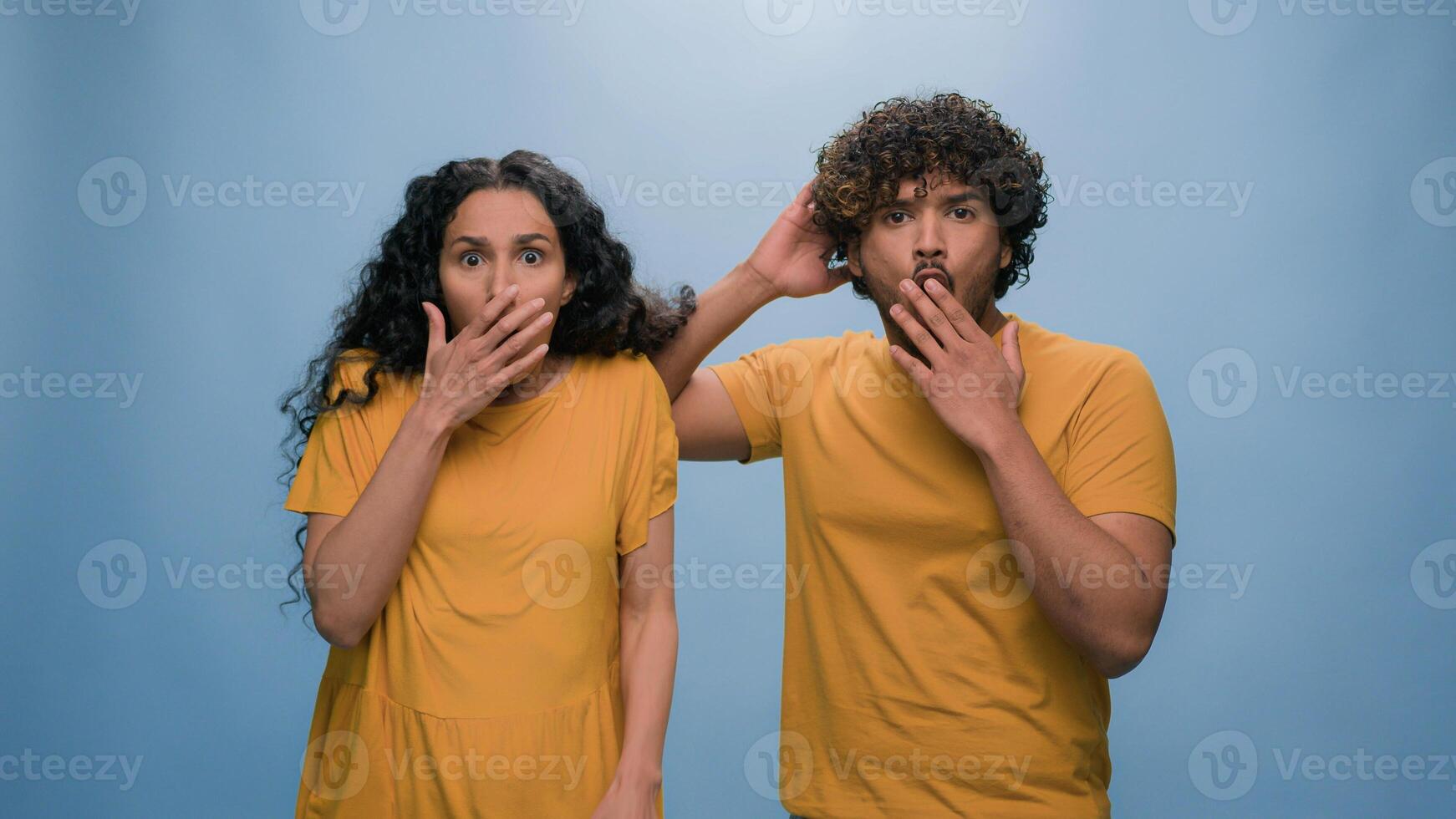 Multiracial shocked scared afraid couple amazed shocked Hispanic woman hiding behind Indian man astonishment wonder scare terrible horror ugly reaction blue studio background shock astonish fright photo