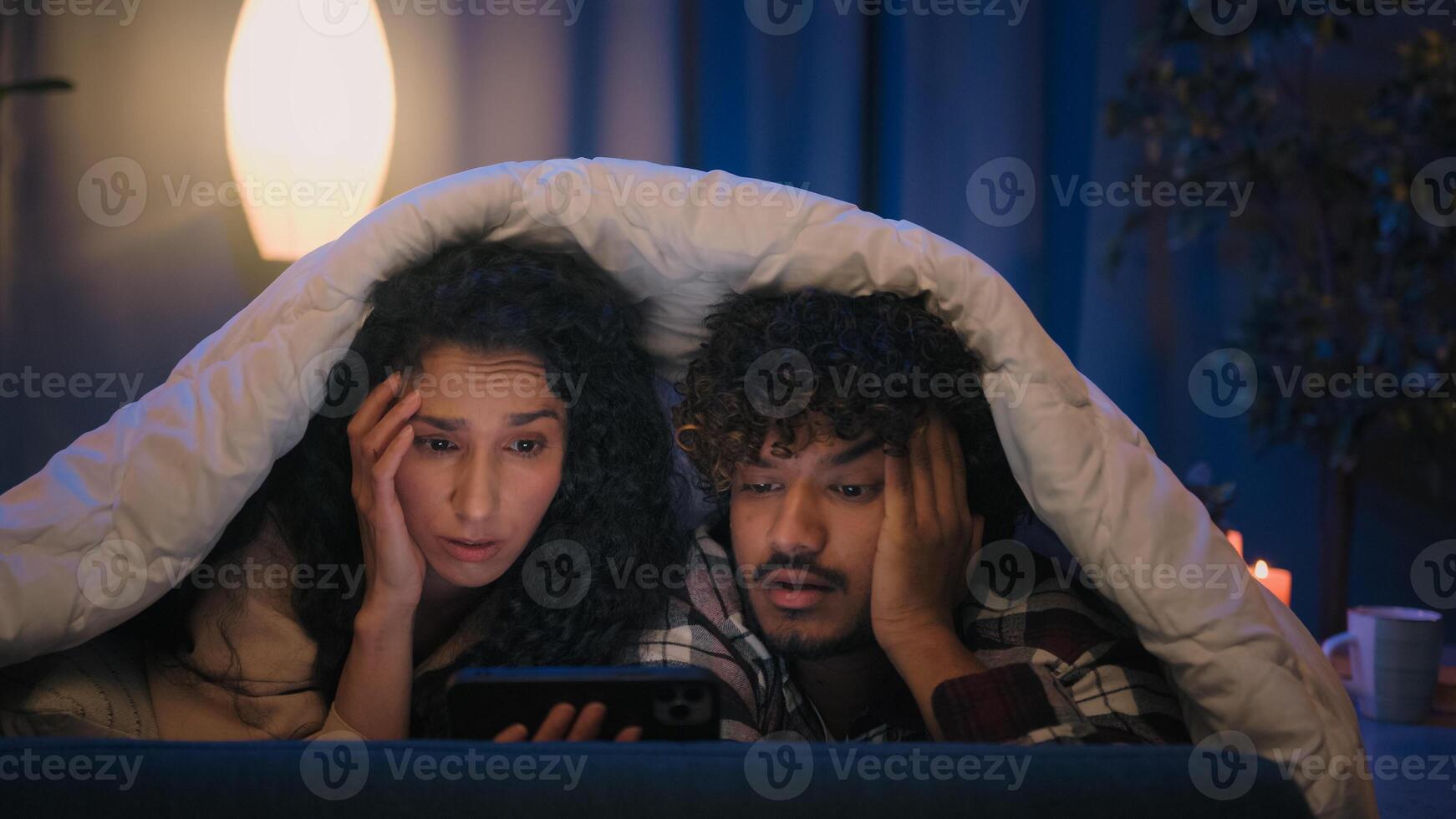 Multiracial sad upset couple man and woman shocked stress disappointed at night home bad news mobile phone lost problem smartphone failure at evening couch under blanket cover with duvet gadget addict photo