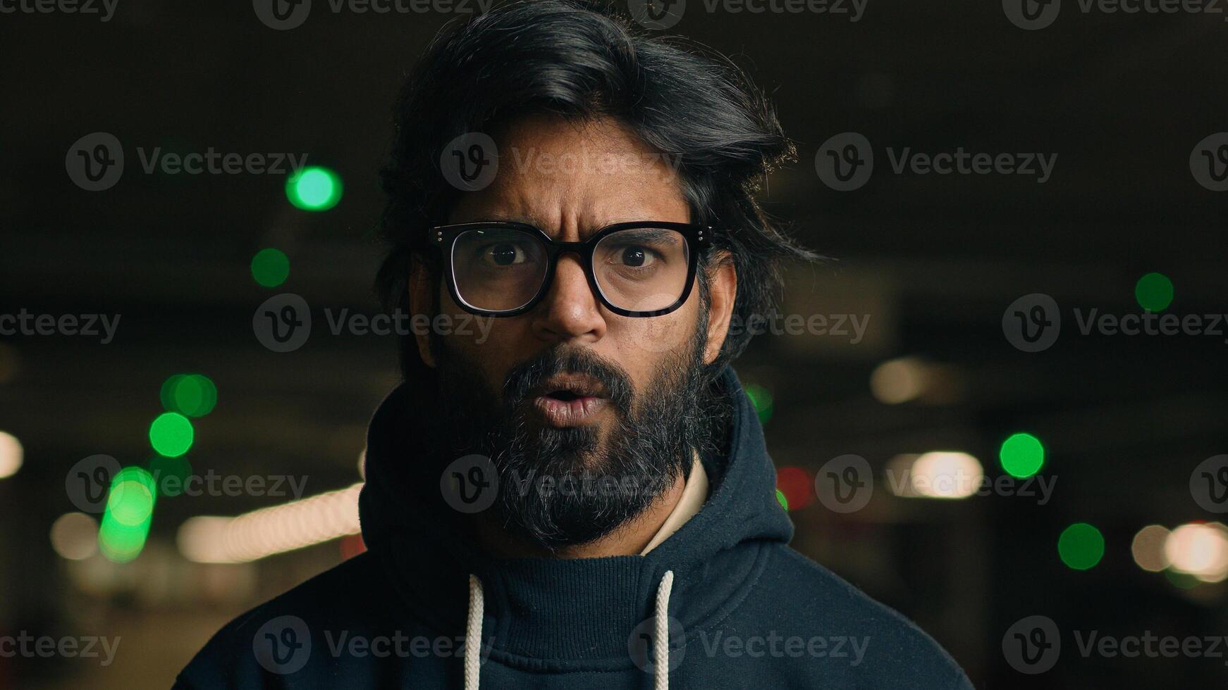 Close up Indian shocked bearded guy in glasses astonished looking at camera at dark underground wonder amazed Arabian man shock fear scared say wow stunned big eyes open mouth bad news at parking lot photo