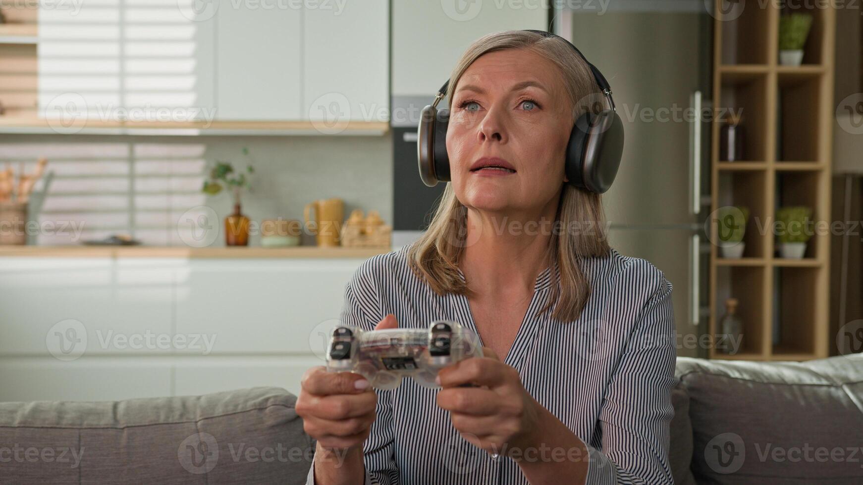 Funny smiling mature caucasian woman playing game console in headphones using joystick controller at home on couch elderly female play computer games older people and modern technology concept photo