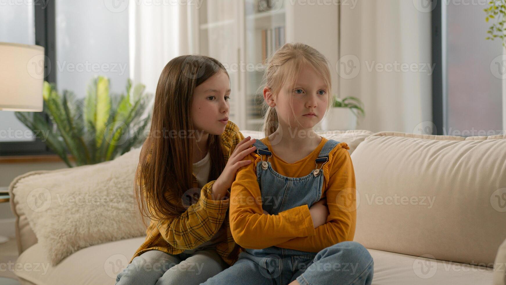 Two little Caucasian kids children siblings daughters girls argument remorseful sister apologizing hugging asking forgiveness younger sister ignoring feel offended angry family quarrel couch indoors photo