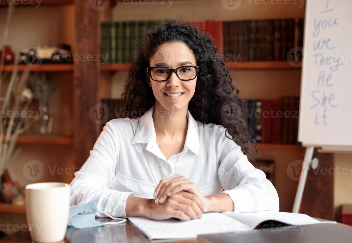 Confident Teacher. Front portrait of smiling curly woman in eyeglasses sitting at desk with bookshelves and board in the blurred background. Casual woman in white shirt posing and looking at camera photo