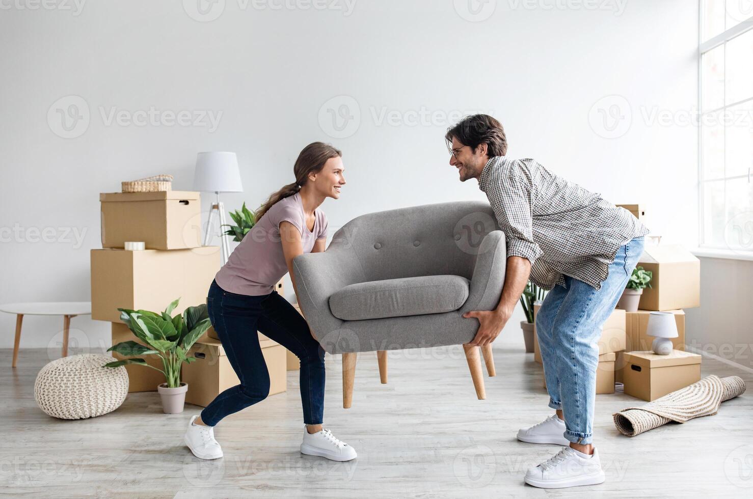 Smiling caucasian millennial wife and husband carry armchair in room with cardboard boxes. Moving together to new house, buy furniture. Purchases apartment on mortgage and loan, rent real estate photo
