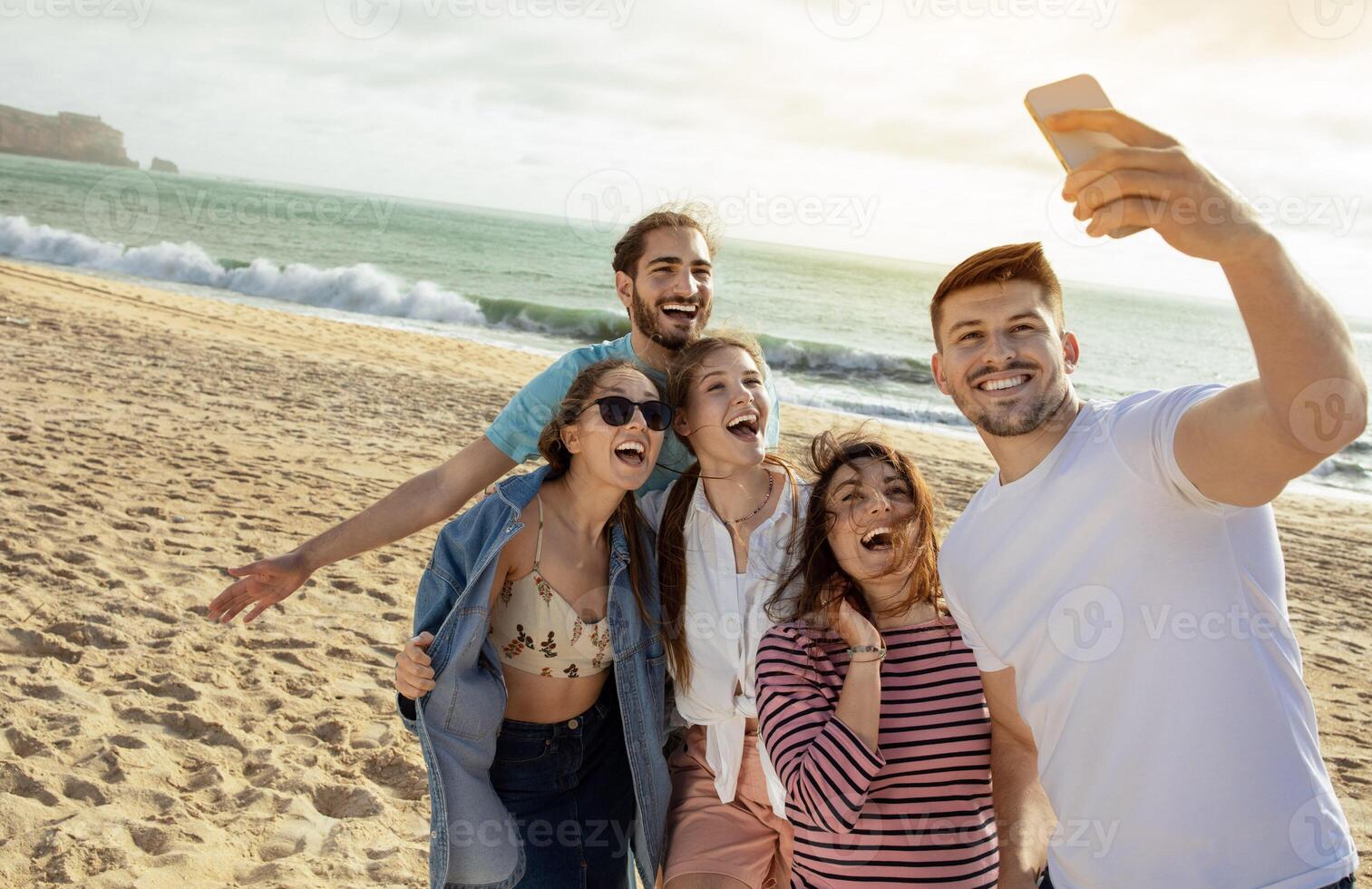 Happy millennial european and arabic friends have fun, taking selfie on smartphone, enjoy free time on ocean beach. App for blog and social network, outdoor party, holiday and trip photo