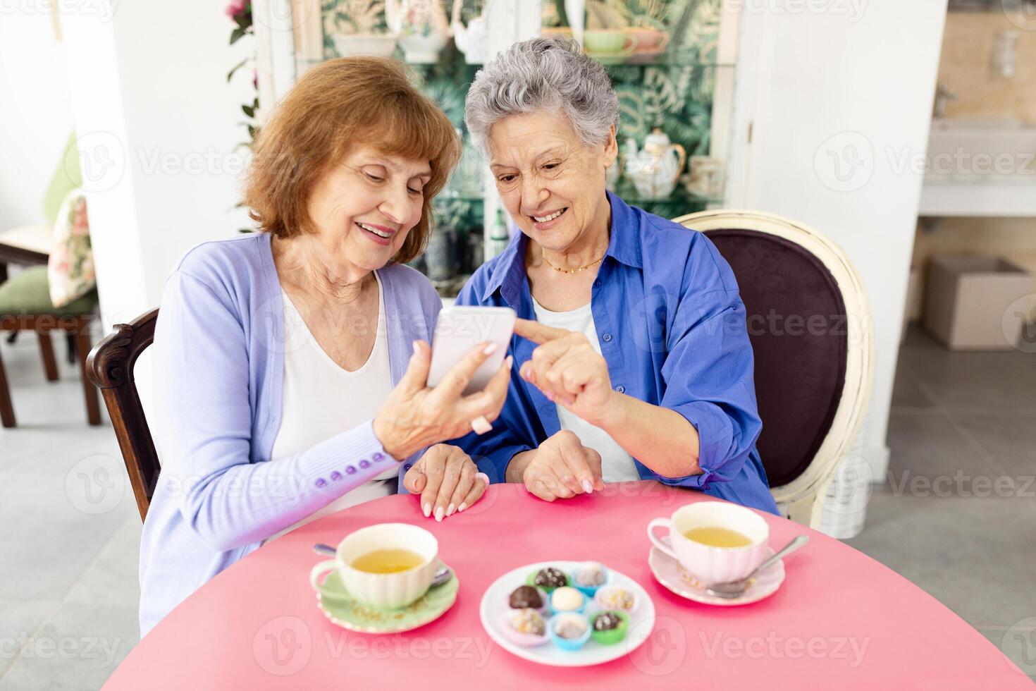 Two elderly women sit together at a round table enjoying dessert and tea. They are sharing laughter while looking at a mobile device. The cafe setting has a cozy ambiance. photo