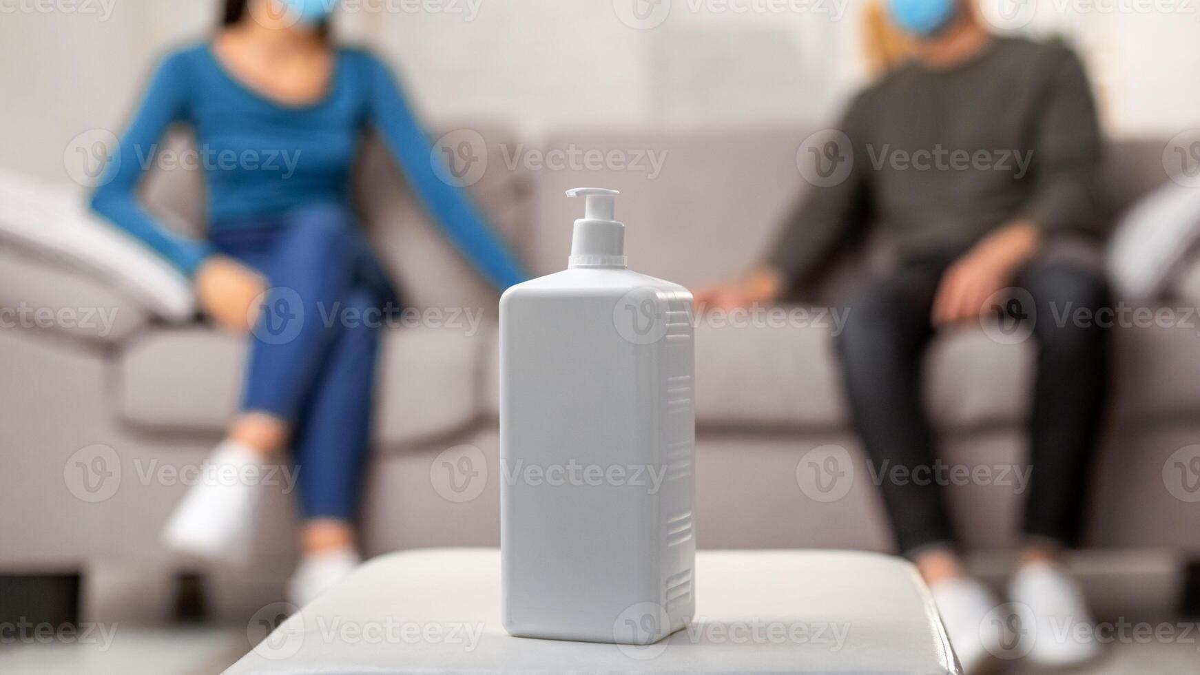 Hygiene Concept. Selective focus on sanitizer bottle on table, young couple in masks sitting in the background photo