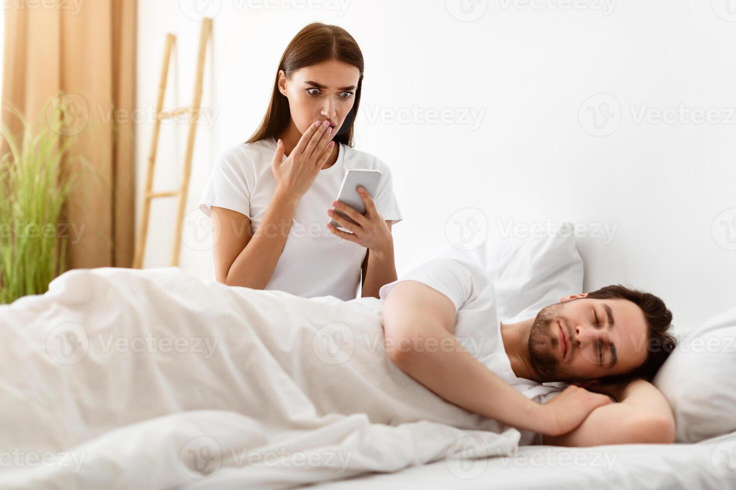 Infidelity. Shocked Wife Reading Message On Cheating Husband's Smartphone While Man Sleeping In Bedroom At Home. Jealousy And Unfaithfulness In Relationship Concept photo