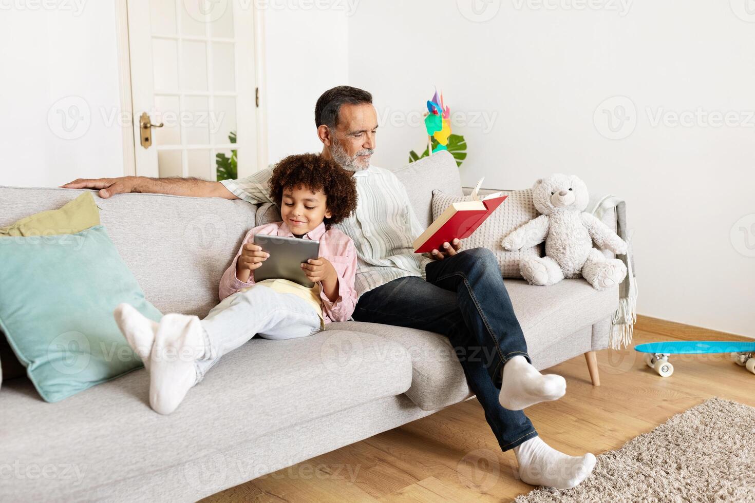 A grandfather and his grandson enjoy a cozy afternoon at home. The boy plays on a tablet while the elder reads a book, showcasing their bond and love for each other. photo
