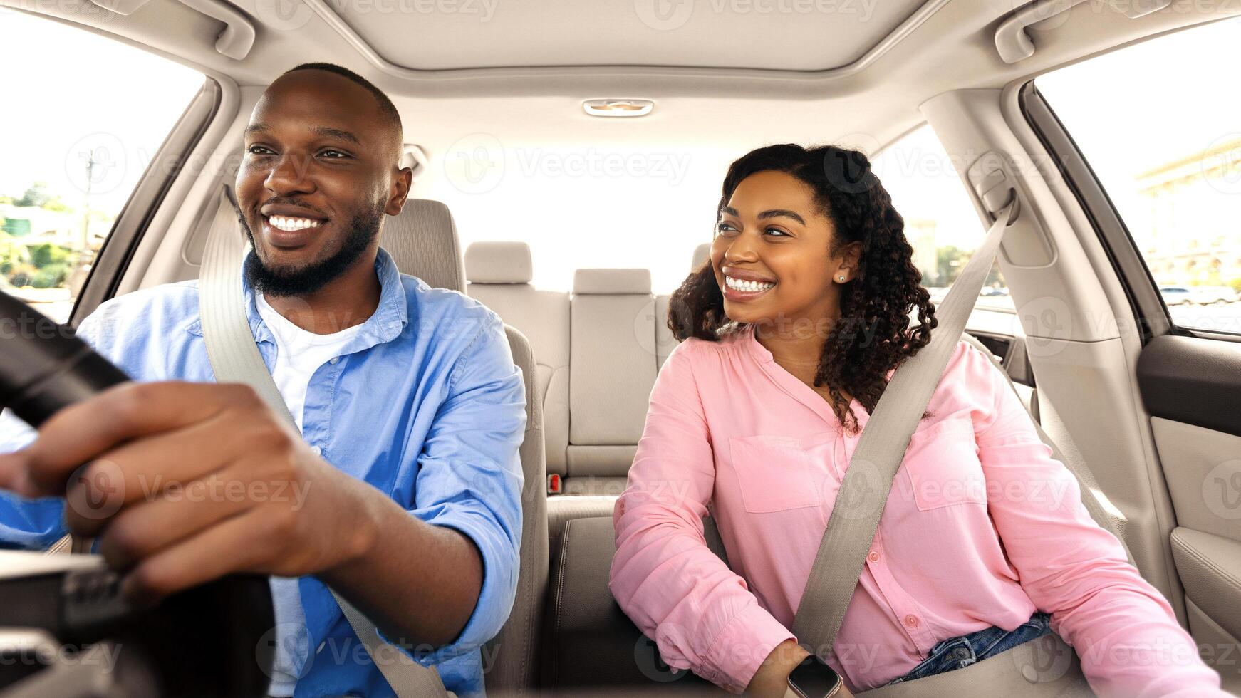 Enjoying Travel Concept. Smiling African American man driving modern luxury car, happy young woman sitting on the front passenger seat, looking on the driver, family going on roadtrip, windshield view photo