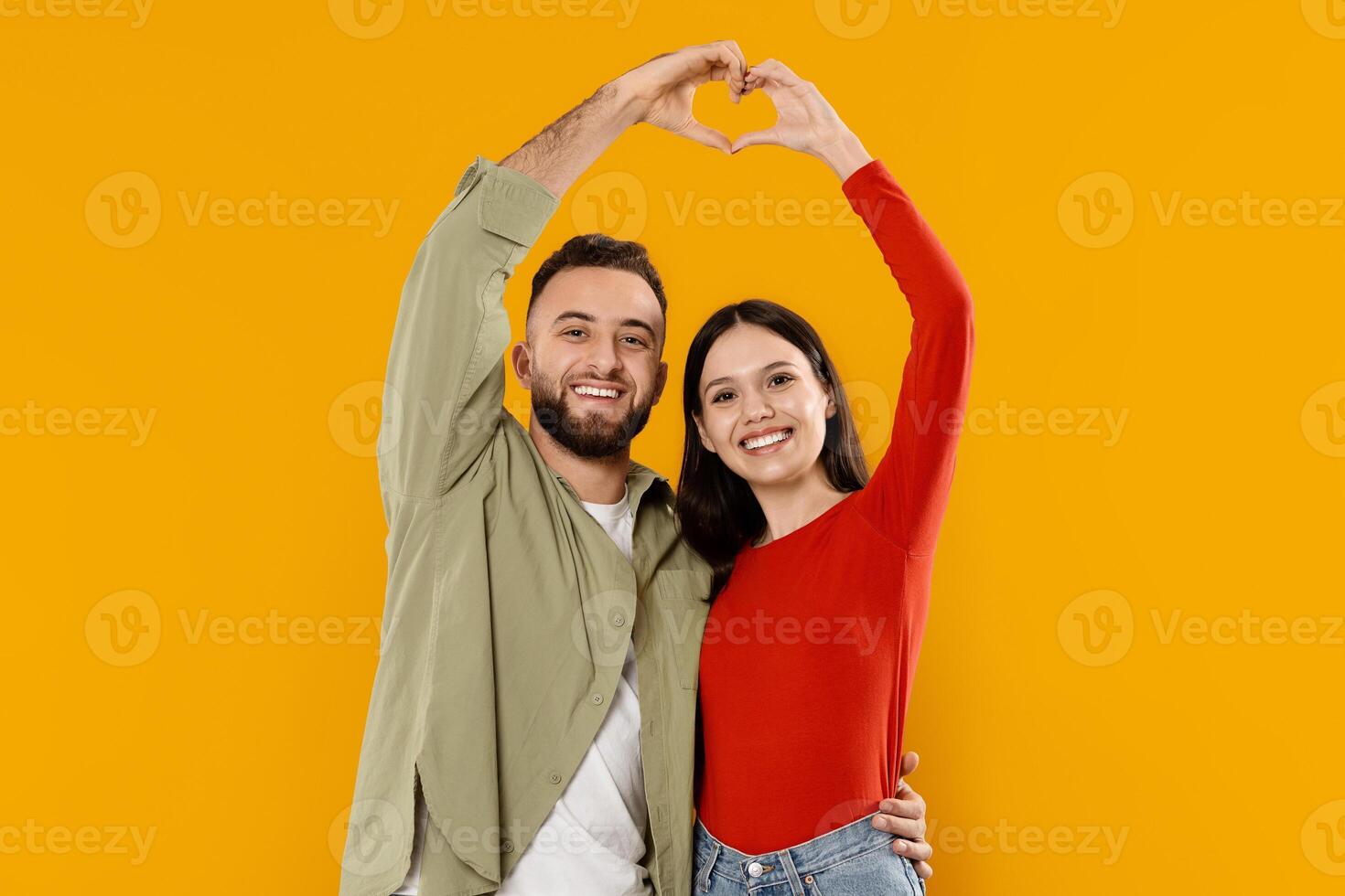 A young married couple stands together, smiling brightly. They form a heart shape with their hands, expressing love and connection, set against a vivid yellow background. photo