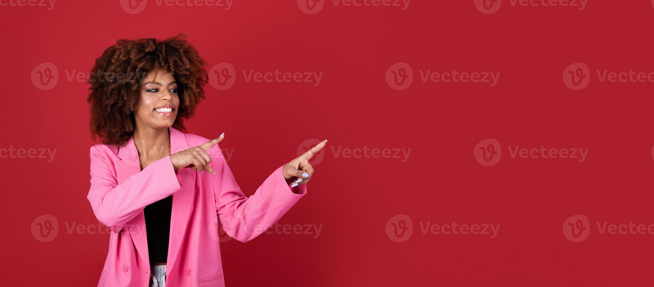 A young African American woman dressed in a stylish pink blazer excitedly points towards copy space against a bold red background, exuding confidence and positivity. photo