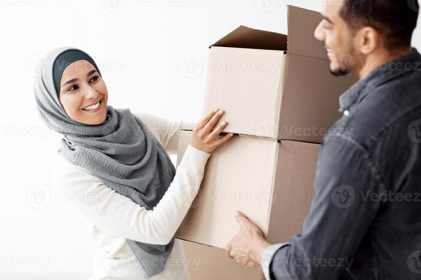 Happy lady in hijab giving her husband boxes with their stuff, moving to new apartment, closeup. Cheerful loving arab family having relocation day, packing belongings into paper boxes photo