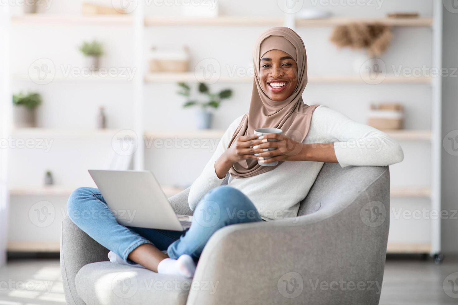 Domestic Leisure. Cheerful Black Muslim Lady In Hijab Relaxing At Home With Laptop Computer And Coffee, Happy Islamic Woman Browsing Internet And Enjoying Hot Drink While Resting In Living Room photo