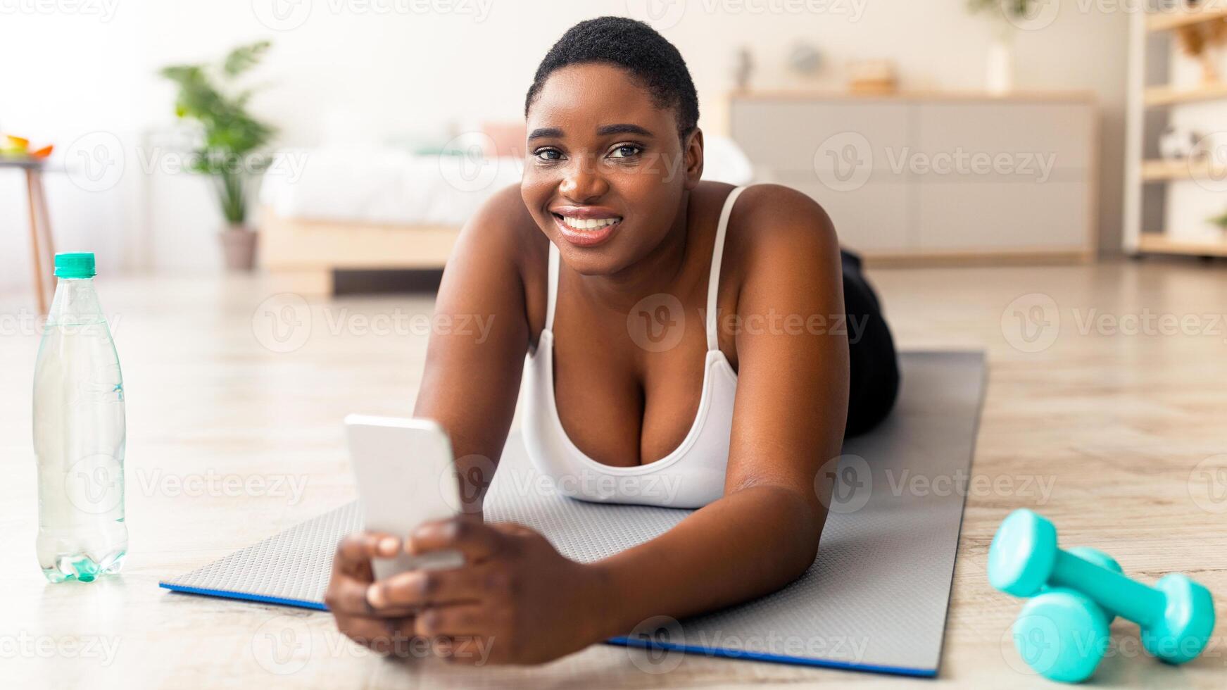 Plump black woman with bottle of water and smartphone browsing internet, checking social network at home. Overweight African American lady having break from domestic sports, using cellphone photo