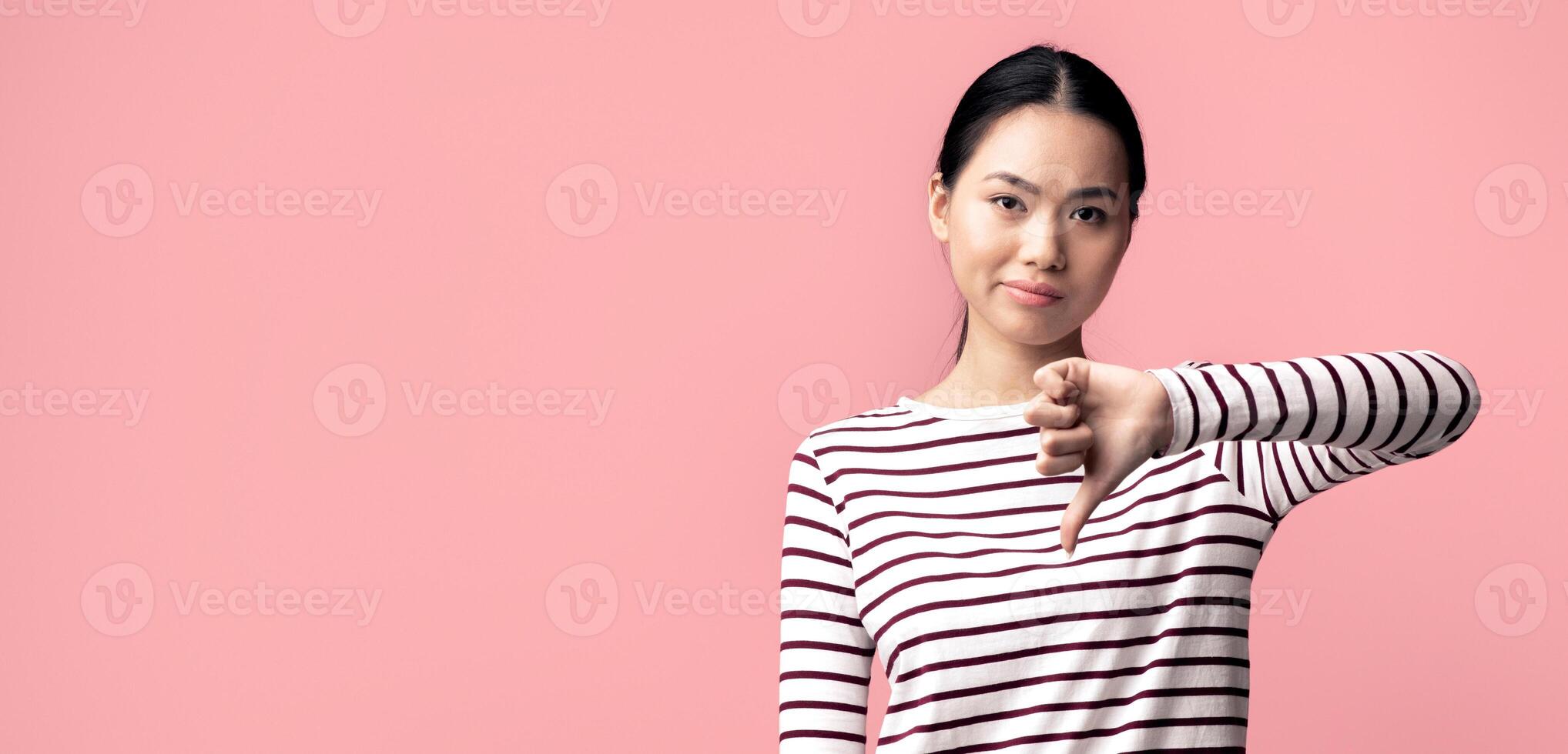 Dislike. Disappointed Asian Female Showing Thumb Down Gesture At Camera, Unhappy Young Korean Woman Expressing Disapproval With mean Face Expression, Standing Over Pink Studio Background, Copy Space photo