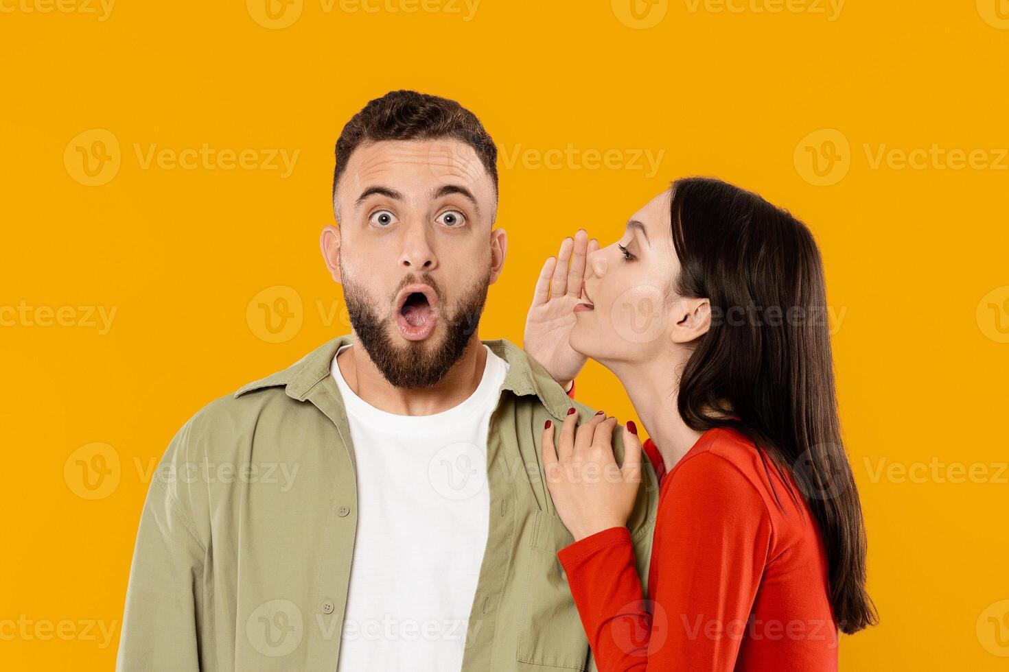 A young man and woman engage in a playful moment, as the woman whispers a secret in the man's ear, who shows surprise. They express affection and joy against a vibrant yellow background. photo