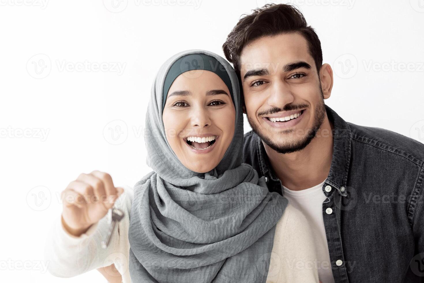 Emotional young lady in hijab showing key from new house, standing by her husband, copy space. Happy muslim family embracing in their brand new apartment, paper boxes with belongings on background photo