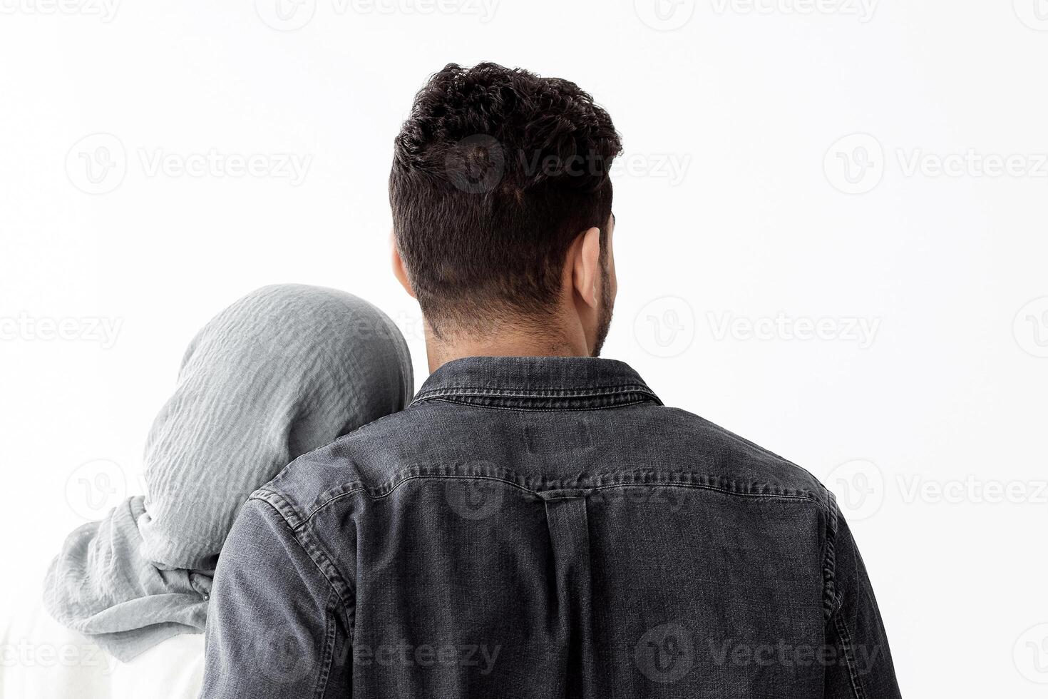 Back view of young muslim family husband and wife cuddling while moving to new house, copy space. Arab man and lady in hijab standing in empty white room, looking at paper boxes and hugging photo