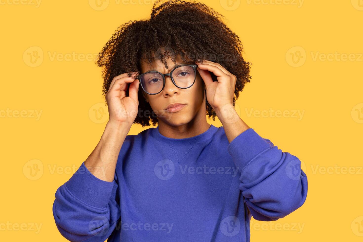 A black guy adjusts his large glasses while wearing a blue sweater. His curly hair frames his face as he engages with the camera, showcasing a modern and confident style. photo