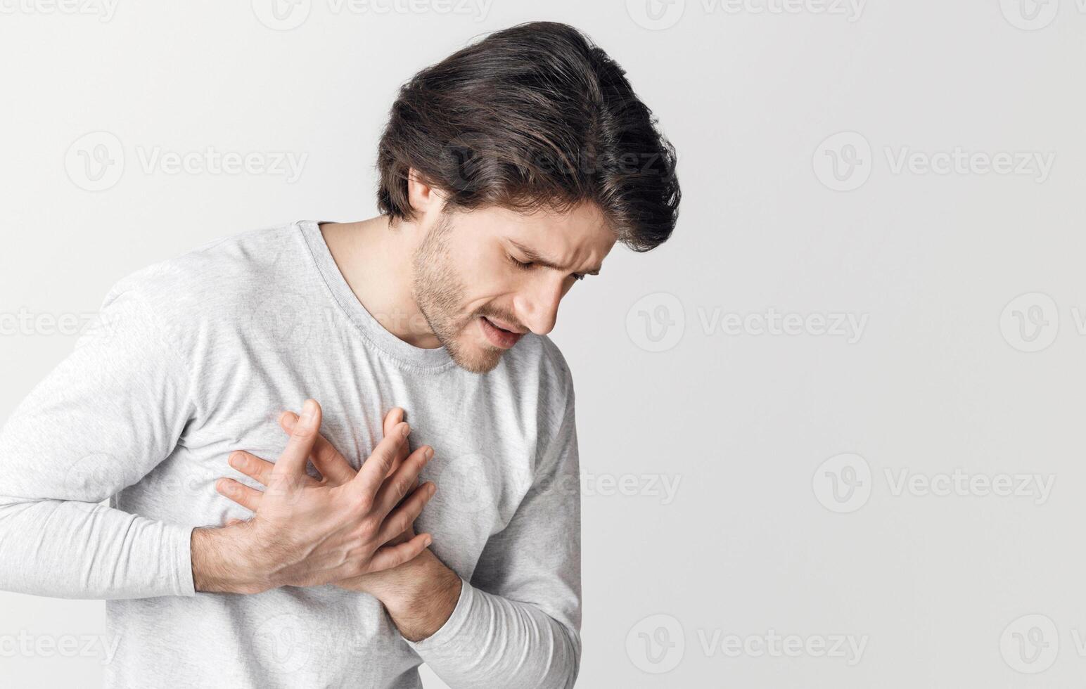 Heart attack at young age. Man presses his hands to chest and facial expression pain, isolated on gray background, studio shot photo