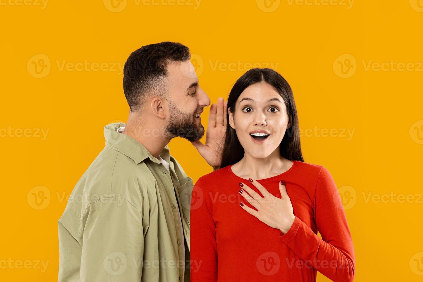 A young couple expresses affection as the man whispers to the woman, who displays surprise and delight. Their connection shines brightly against a bold yellow backdrop. photo
