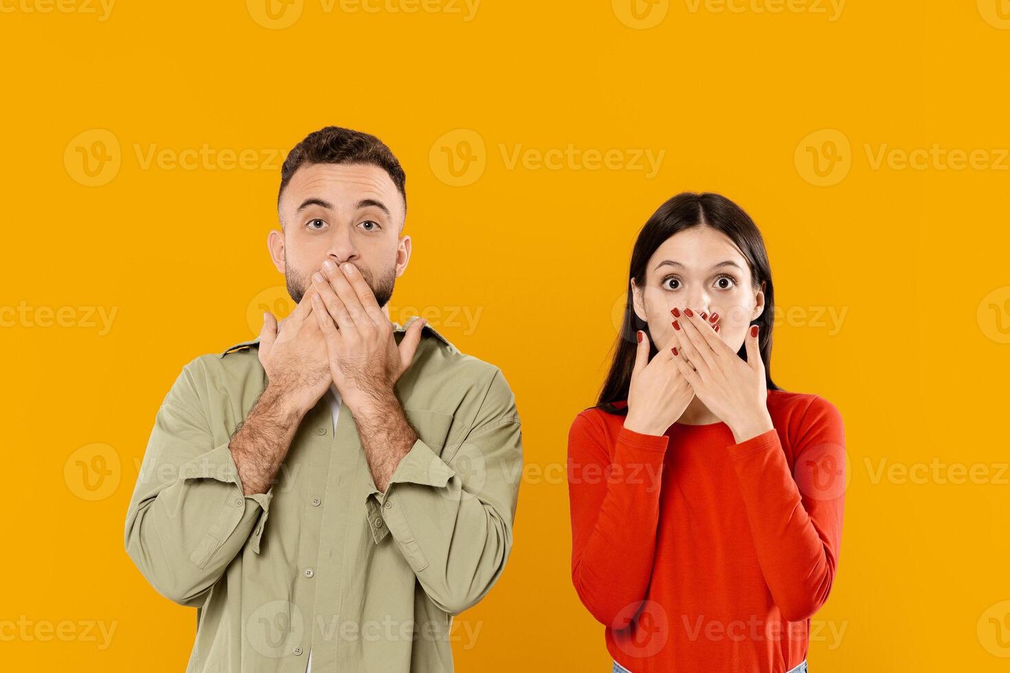 A young couple displays playful surprise, covering their mouths with hands. Set against a bright yellow background, their lively expressions convey fun and affection in their relationship. photo