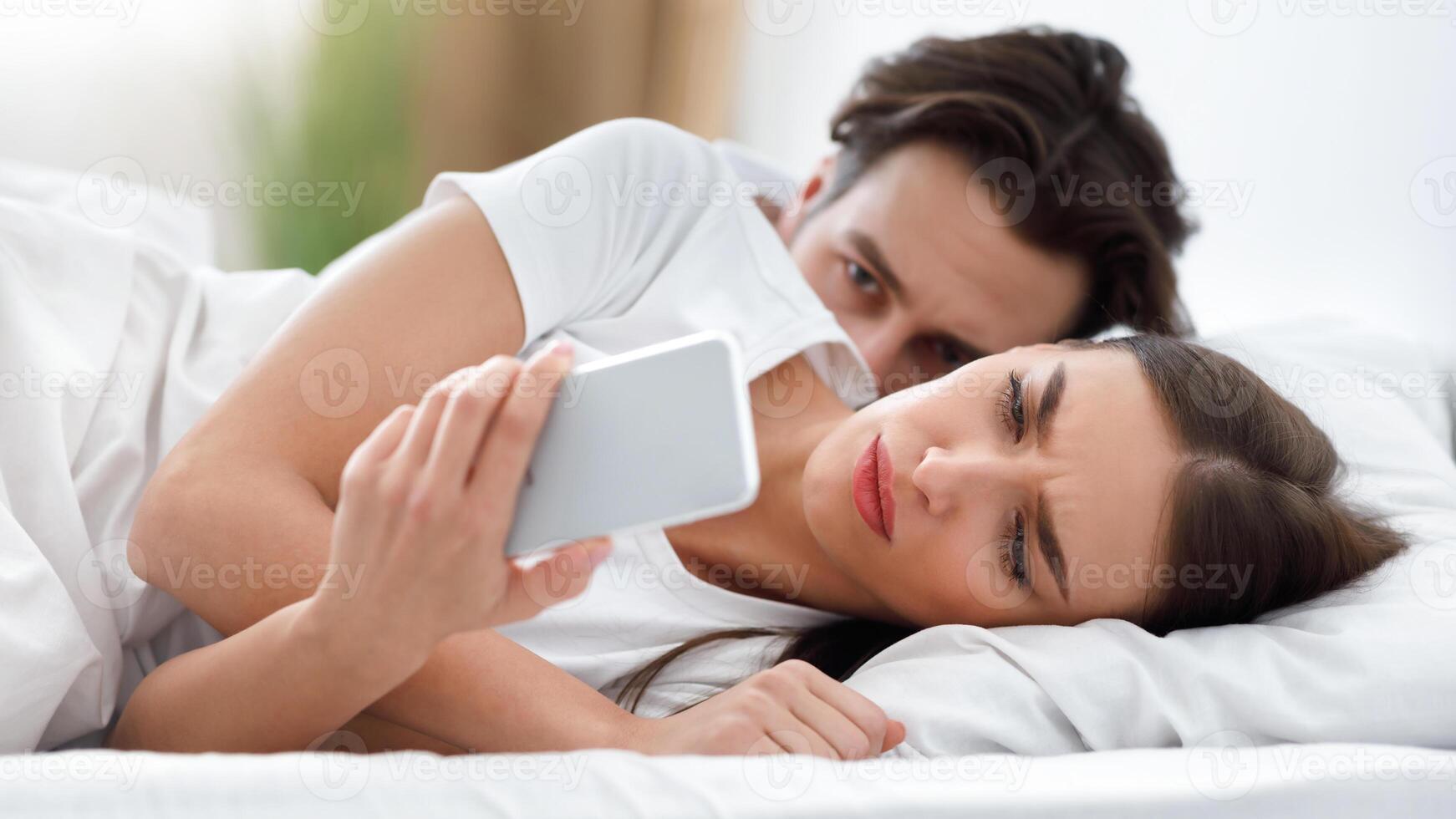 Jealousy In Marriage. Jealous Husband Reading Unfaithful Wife's Messages While She Texting On Cellphone Lying In Bed In Bedroom At Home. Man Suspecting Infidelity In Relationship. Selective Focus photo