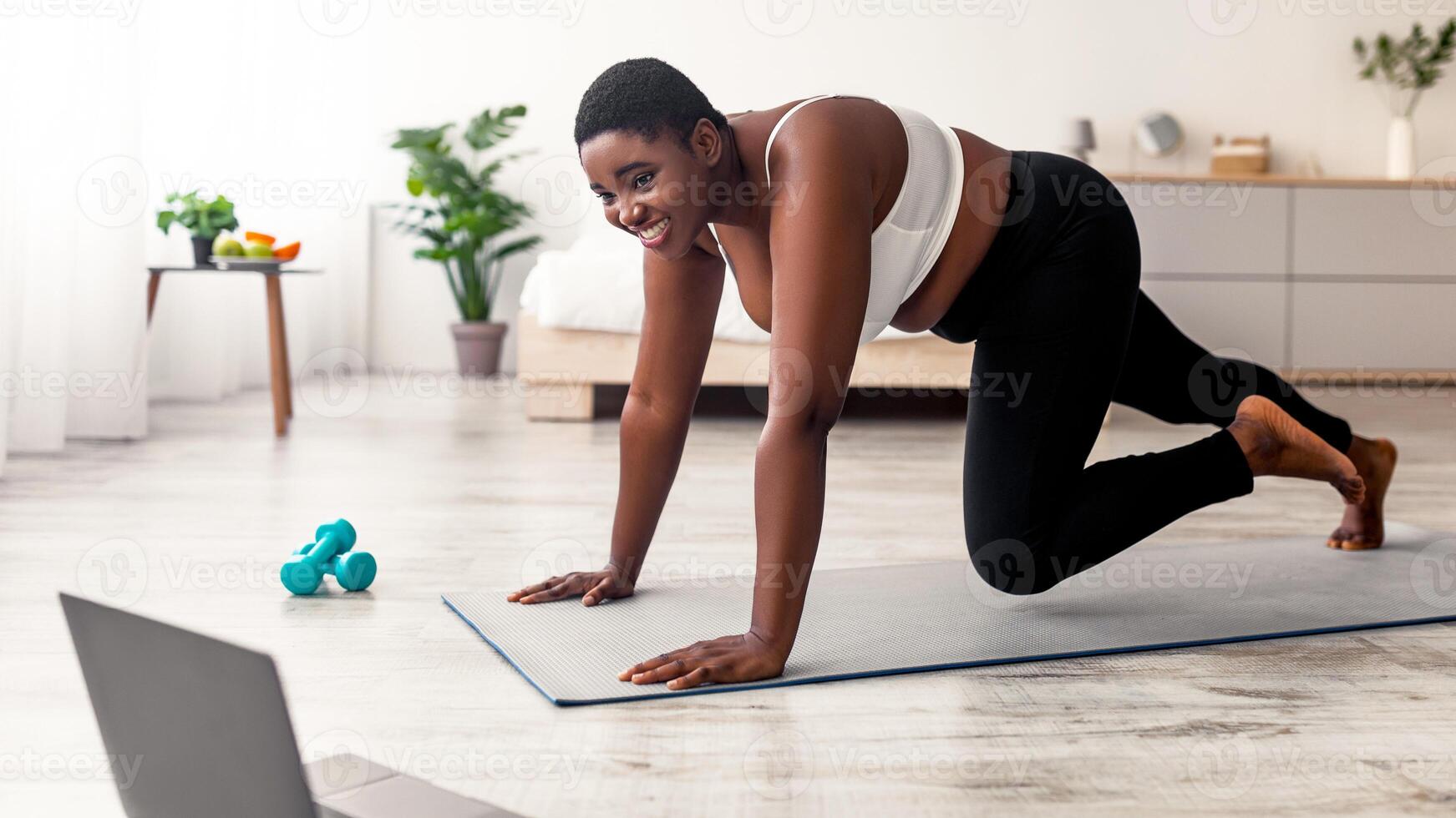 Online bodyweight exercises. Plus size Afro woman having strength workout, following web tutorial on laptop, running with hands on floor at home. Curvy black lady making weight loss training photo