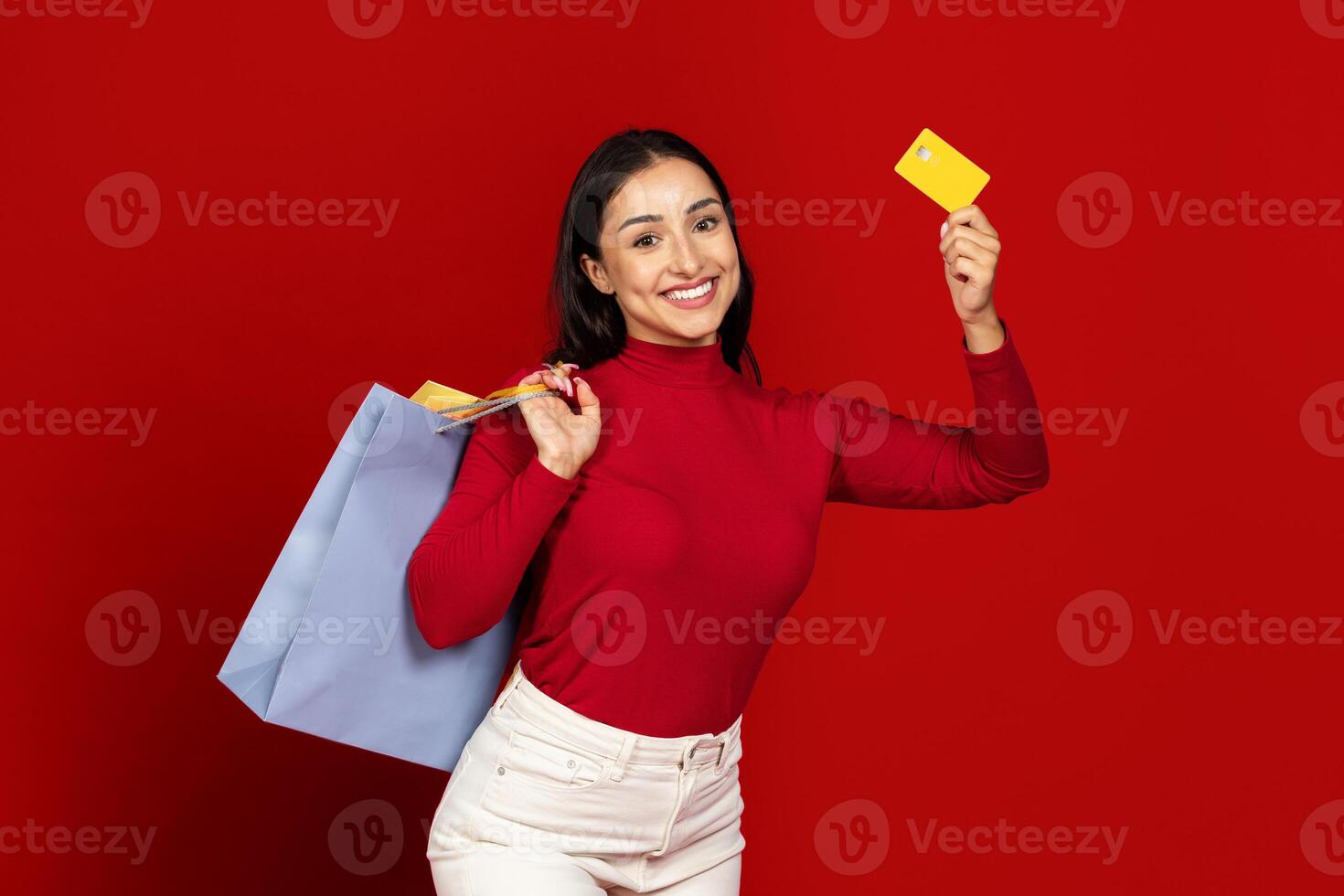 A young Caucasian woman stands cheerfully against a vivid red backdrop, holding a shopping bag and a credit card. photo