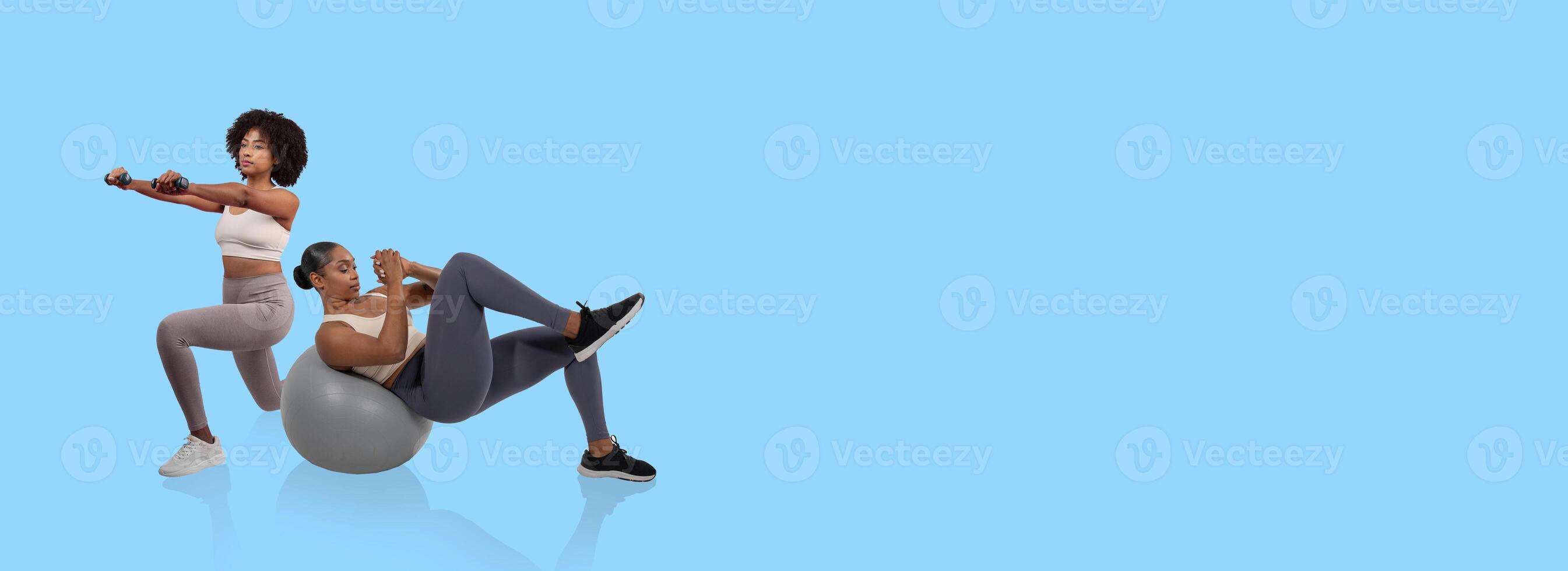 Two women are actively exercising in a fitness studio. One performs a squat with dumbbells while the other completes a crunch on a stability ball, both focused on their workout. photo