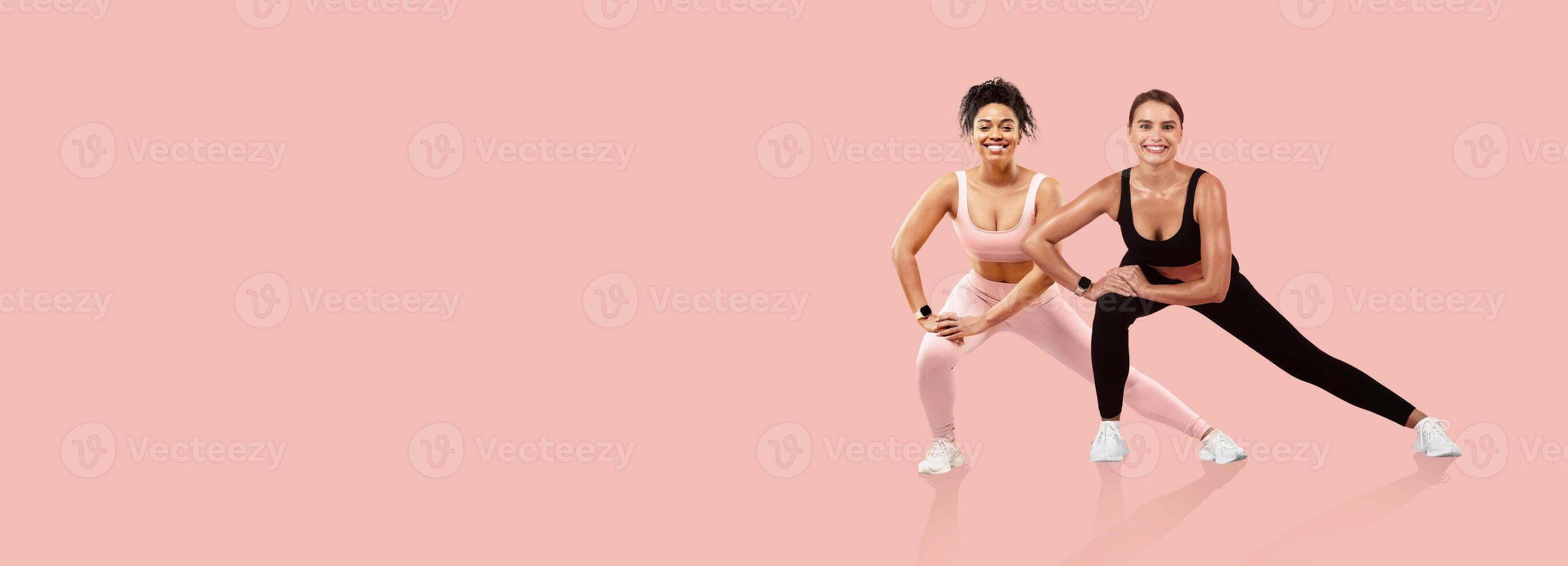 Two women engage in a workout routine, showcasing teamwork and motivation. They wear vibrant activewear and demonstrate an exercise on a pastel pink background, emphasizing fitness. photo