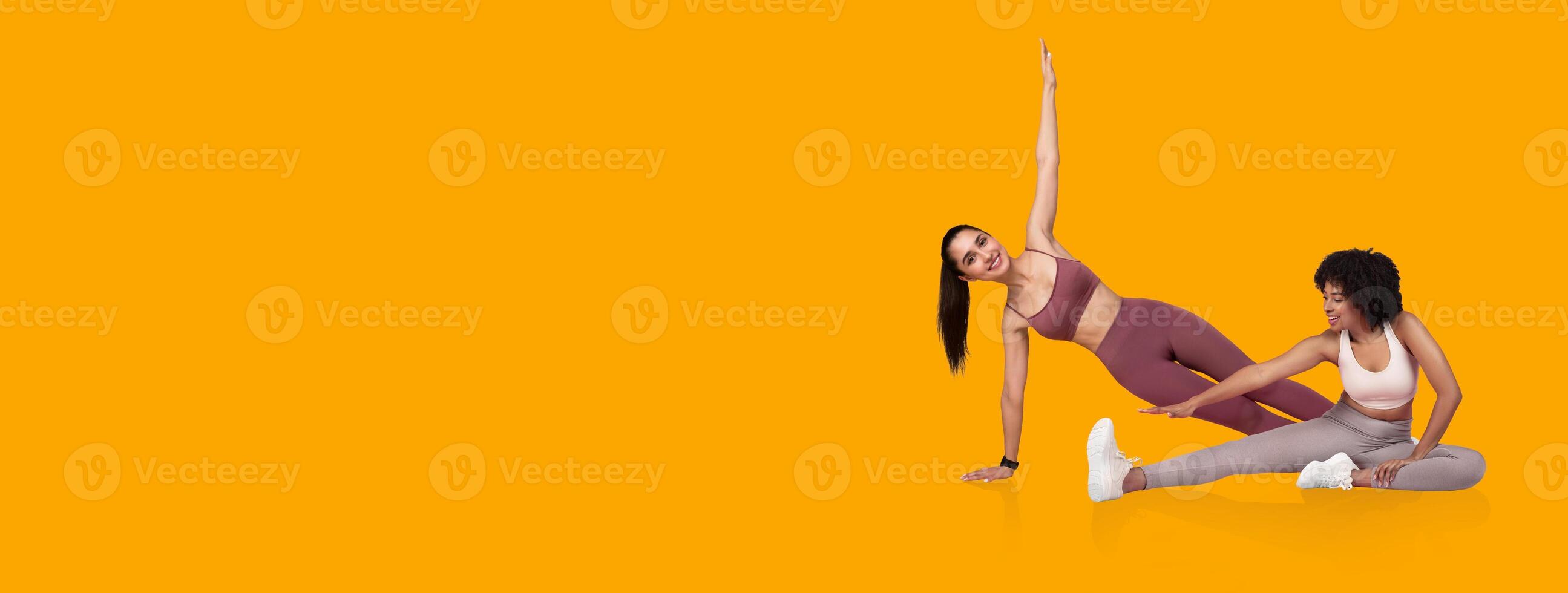 Two women are performing yoga poses on a vibrant yellow background. One woman stretches sideways while the other supports her in a dynamic fitness activity. photo