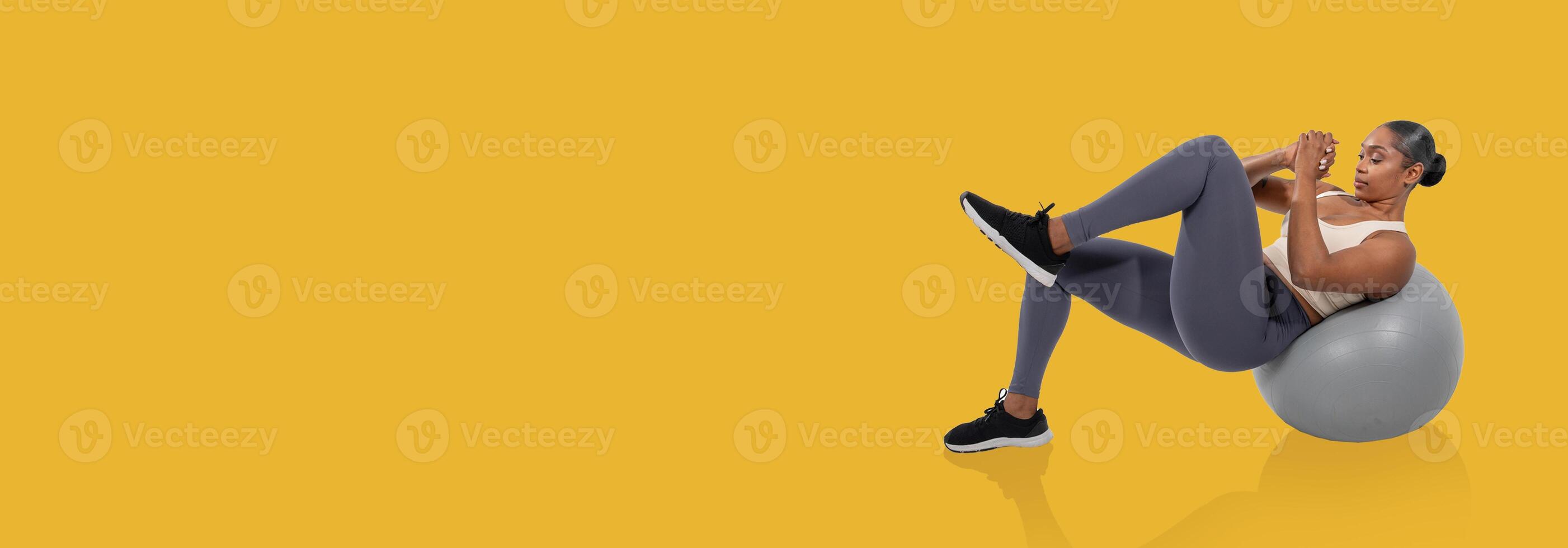 A woman is engaging in a core workout on a stability ball, enhancing her fitness routine. She wears athletic clothing and focuses on her form, set against a bright yellow background. photo