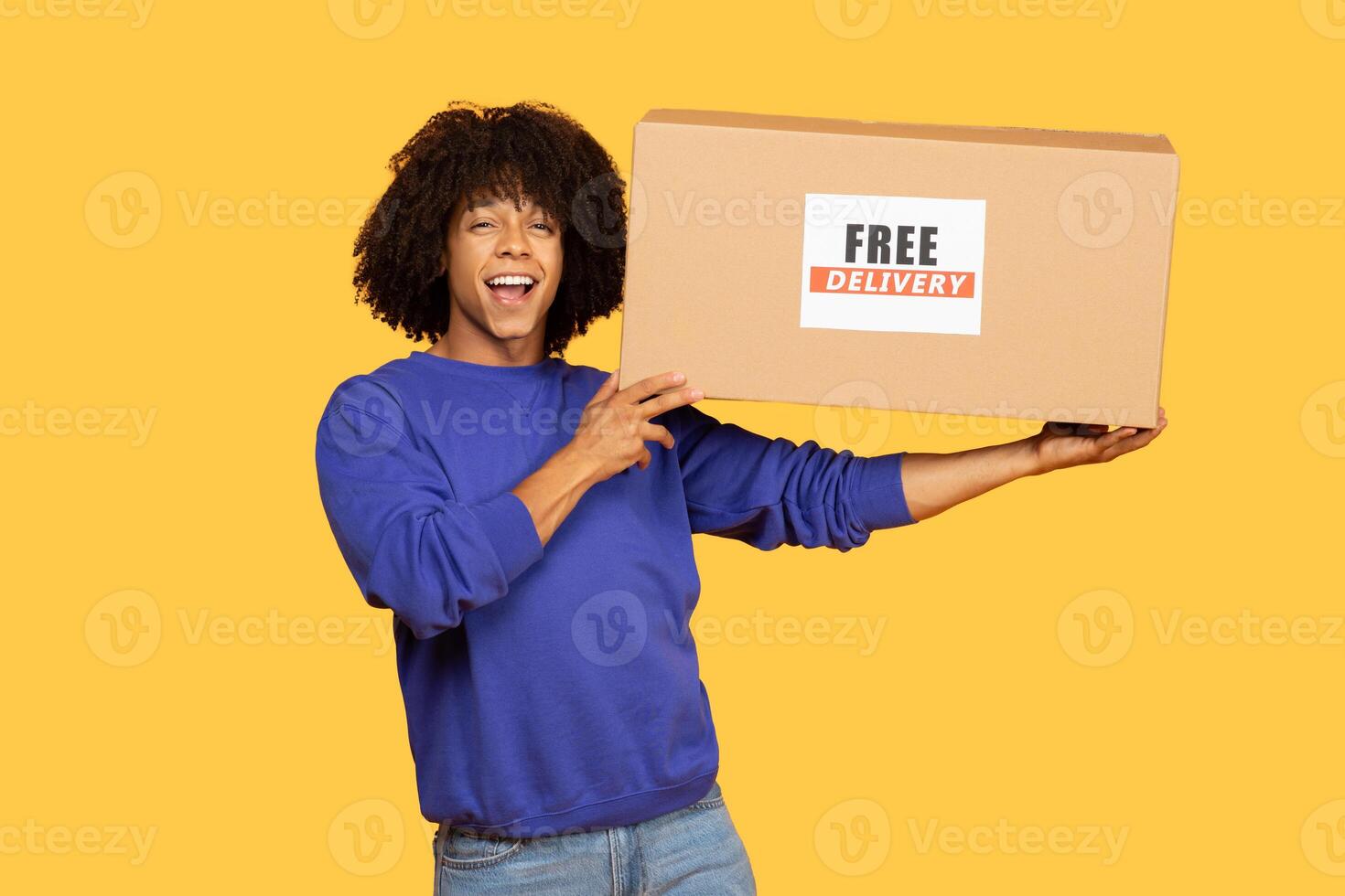 A Black man enjoying a moment of excitement holds a large cardboard box labeled free delivery. The background is bright yellow, creating a lively atmosphere as he smiles and engages with the camera. photo