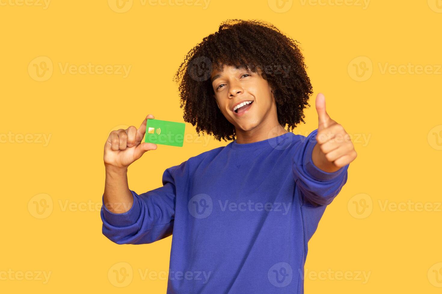 A cheerful young man with curly hair holds a green card in one hand while giving a thumbs up with the other. He stands against a vibrant yellow backdrop, expressing excitement and confidence. photo