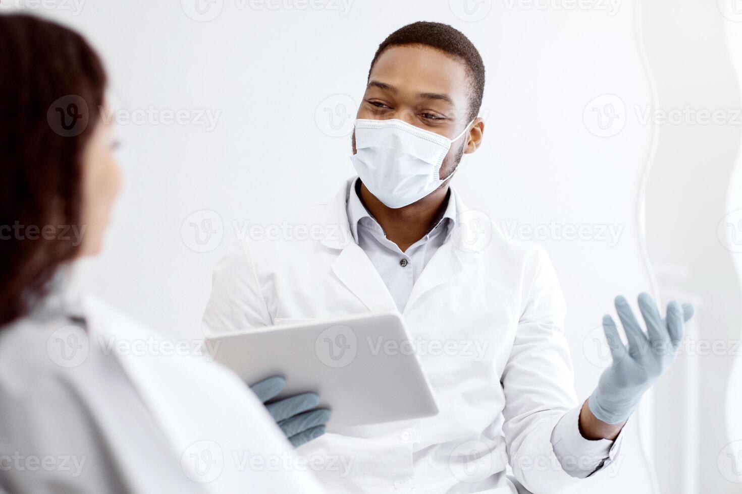 Young Black Dentist Doctor With Digital Tablet In Hands Consulting Patient In Clinic, African American Stomatologist Explaining Teeth Treatment Process To Customer During Check Up In Dental Cabinet photo