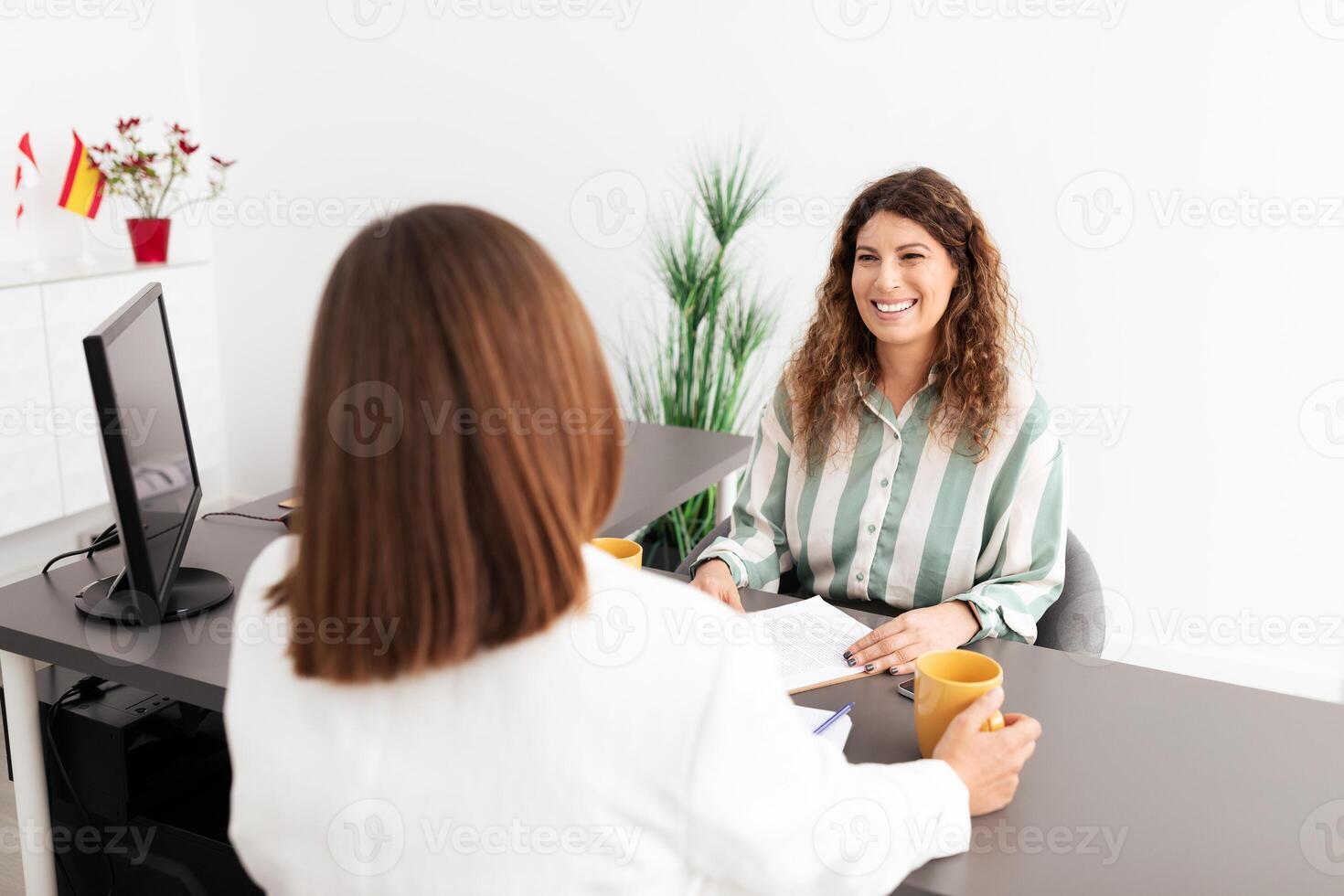 Two Caucasian women are discussing work matters in a sleek office environment. One woman holds a coffee cup while the other smiles, showcasing collaboration and professionalism. photo